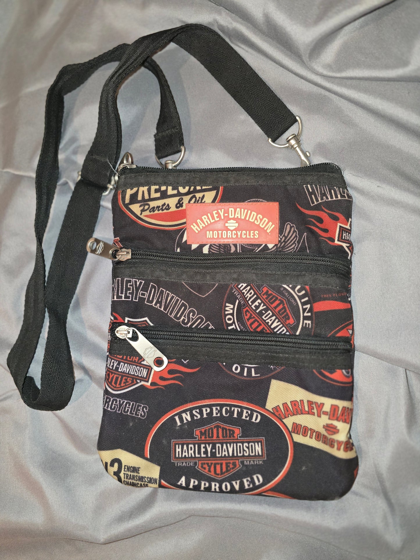 Sling this pre-owned Harley-Davidson® Women's X-Body Oil Print Crossbody Bag over your shoulder and show off your edgy side. Made with zipper pull logos and quality craftsmanship, this bag is both stylish and functional. Great for any occasion, this bag is sure to make a statement! Noted the strap on the back is coming loose, but feels secure.
