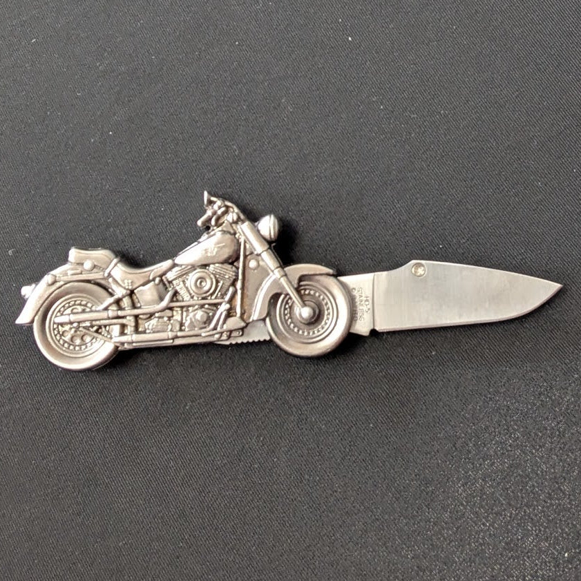A silver folding pocket knife with a detailed motorcycle design on the blade, resting on a dark gray surface.