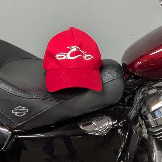 A red baseball cap with a white embroidered motorcycle graphic sits on the black seat of a red and black Harley-Davidson motorcycle. The motorcycle is parked indoors against a gray wall, with a black jacket hanging on the wall above.