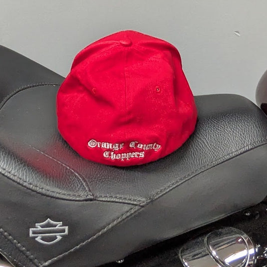 A red baseball cap with white embroidered text sits on the black leather seat of a motorcycle in a garage setting.