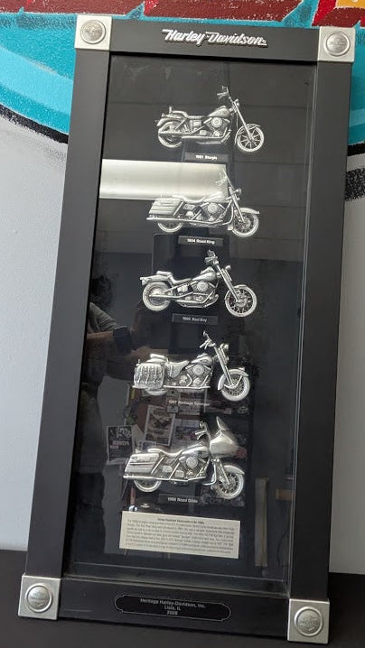 A Harley-Davidson display case featuring five silver motorcycle models inside a black glass cabinet, set against a colorful wall mural.