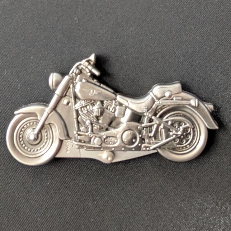 A small silver motorcycle-shaped pin or charm with detailed engine and wheel features, photographed on a dark gray textured background.