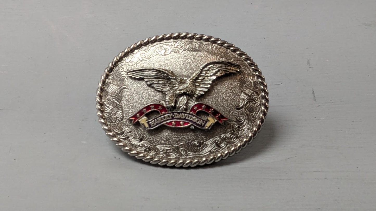 A silver-toned oval metal belt buckle featuring an eagle design with red and gold enamel details, photographed on a plain gray surface.