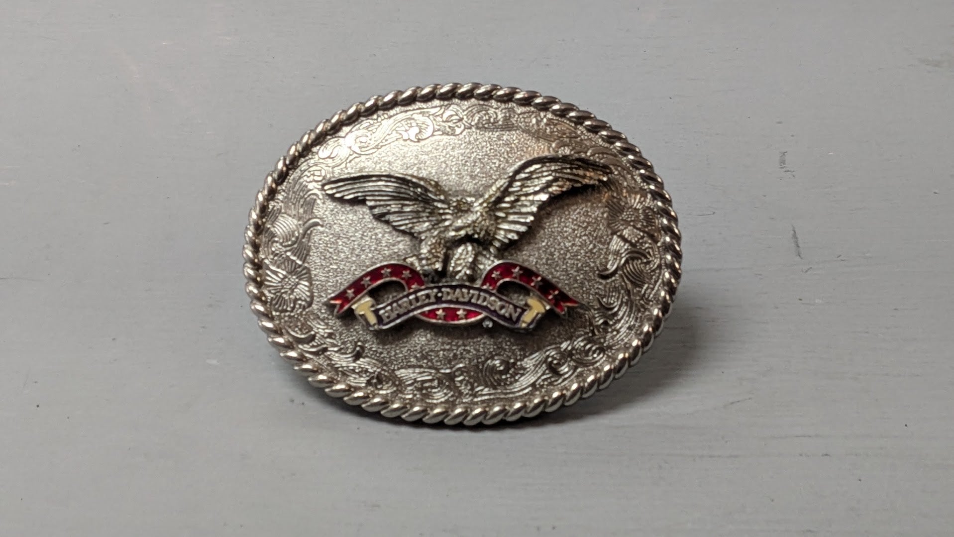 A silver-toned oval metal belt buckle featuring an eagle design with red and gold enamel details, photographed on a plain gray surface.
