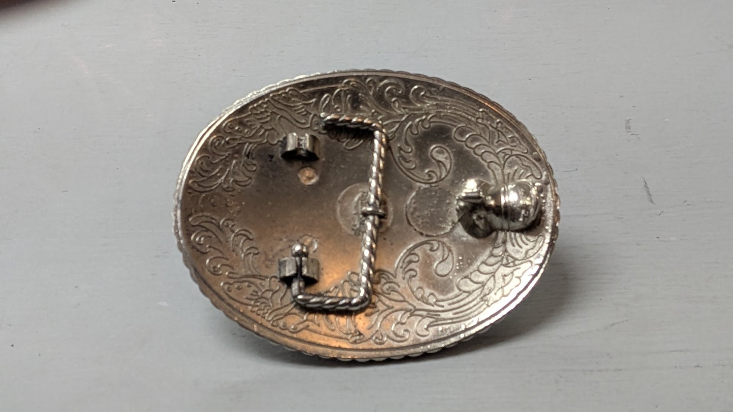 An oval silver-toned metal belt buckle with ornate engraved floral scrollwork and a central twisted rope detail, photographed on a plain light gray surface.