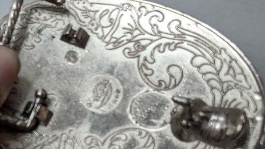 A close-up of a hand holding a silver oval pin or badge with ornate engraved floral patterns and a small horse emblem, set against a plain gray background.