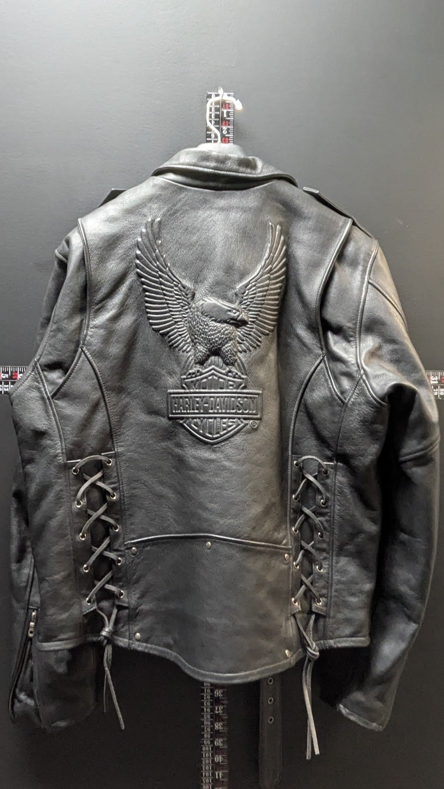 A black leather motorcycle jacket with an embossed eagle and Harley-Davidson bar-and-shield patch on the back, displayed on a hanger against a dark gray wall.
