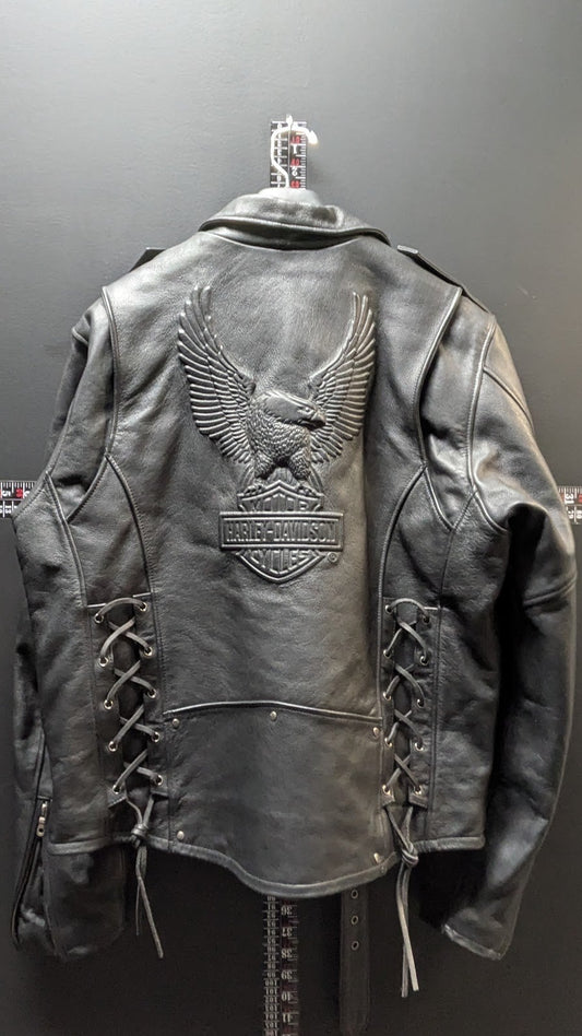 A black leather motorcycle jacket with an embossed eagle and Harley-Davidson bar-and-shield patch on the back, displayed on a hanger against a dark gray wall.