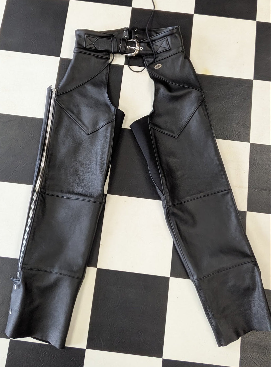 Black leather chaps pants with a matching belt and silver buckle, laid flat on a black and white checkered floor.