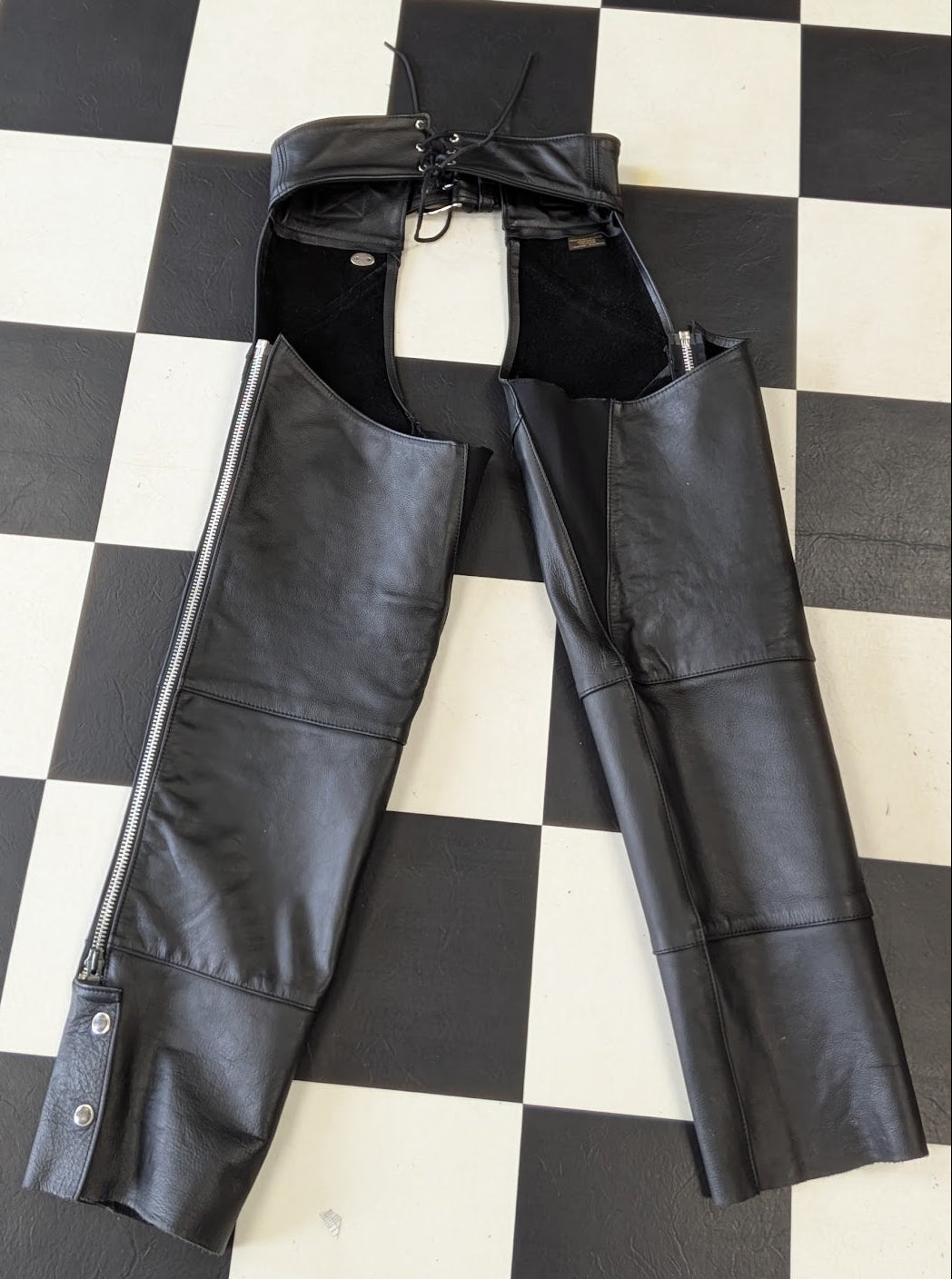 Black leather chaps with a front zipper and adjustable drawstring waist, laid flat on a black and white checkered floor.