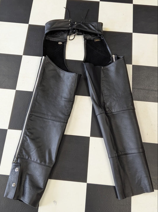 Black leather chaps with a front zipper and adjustable drawstring waist, laid flat on a black and white checkered floor.
