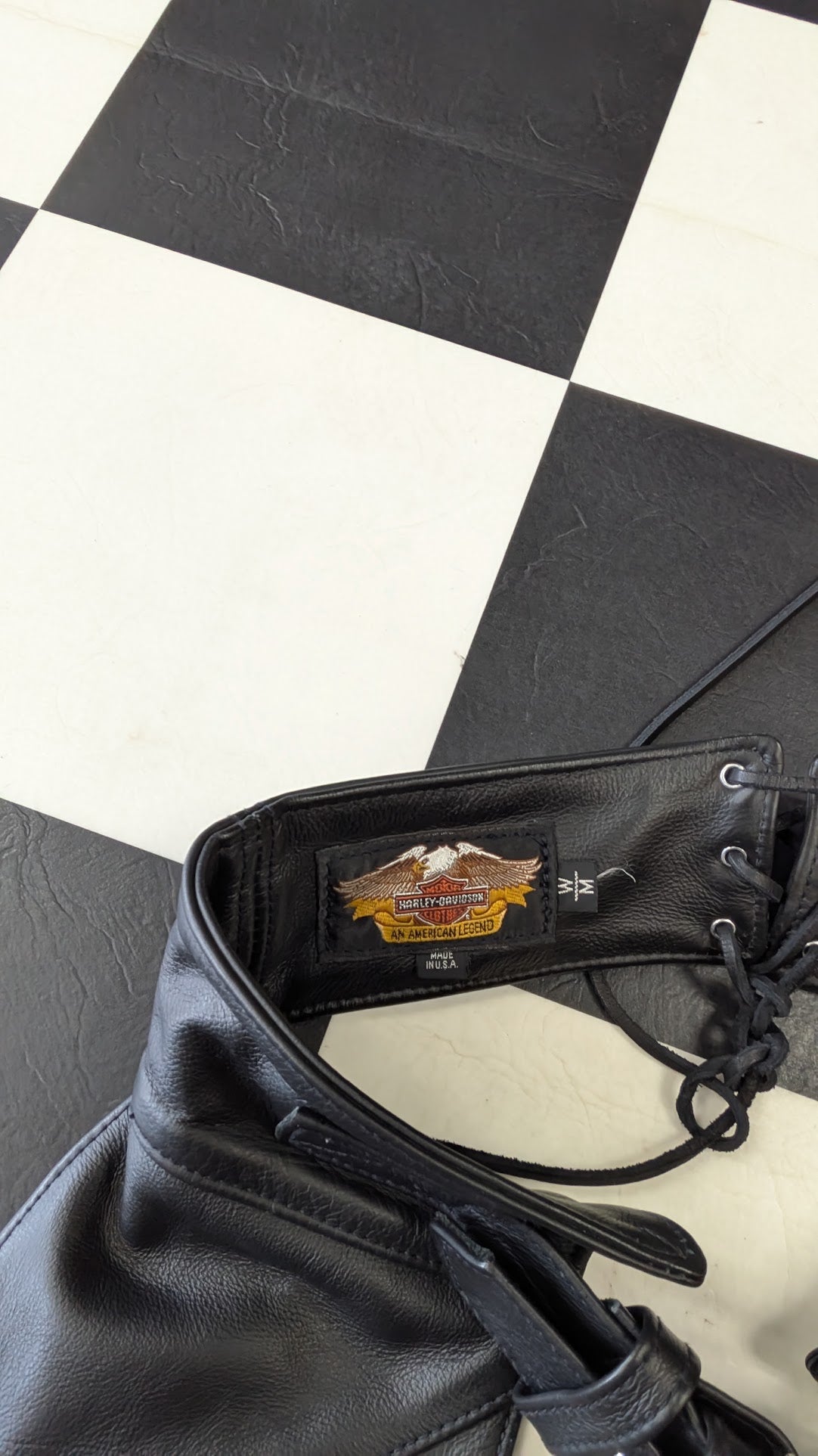 A close-up of a black leather motorcycle boot with a visible Harley-Davidson patch on the collar, resting on a black and white checkered floor.