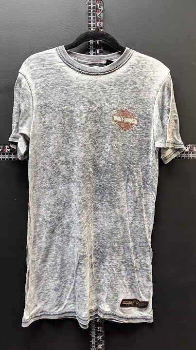 A gray mineral-wash short-sleeve t-shirt with a small brown Harley-Davidson logo on the left chest, hanging on a black hanger against a dark gray wall.