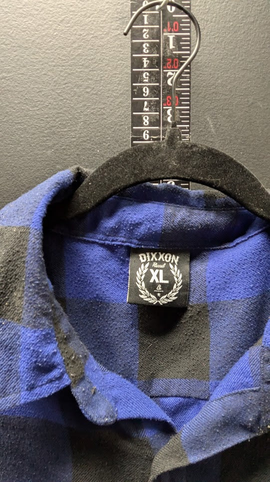 A close-up of a blue and black plaid shirt collar hanging on a black hanger against a gray wall, with a measuring tape visible behind the collar and a visible Dixxon XL neck label.