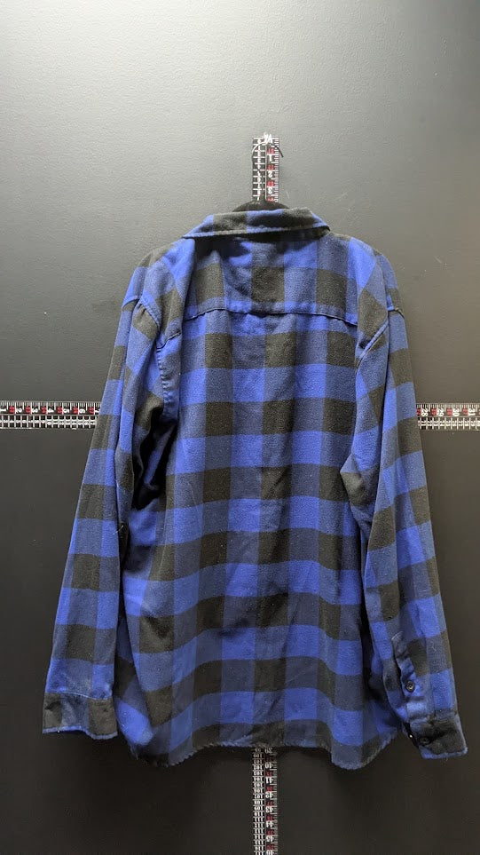 A blue and dark gray plaid flannel shirt hanging on a hook against a dark gray wall, with a measuring tape visible behind it.