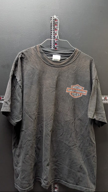 A faded dark gray short-sleeve t-shirt with a distressed wash, featuring a small orange and white Harley-Davidson logo on the left chest, hanging on a metal hanger against a dark gray wall.
