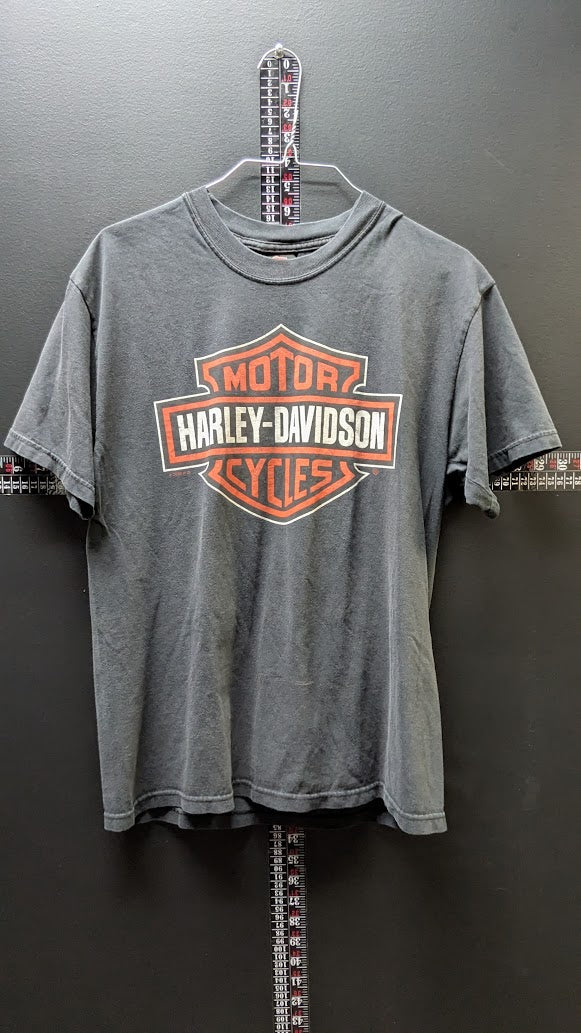 A gray washed Harley-Davidson t-shirt hanging on a hanger against a dark gray wall, featuring the classic orange and white bar-and-shield logo on the chest.