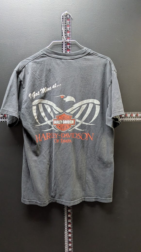 A gray short-sleeve t-shirt hanging on a hanger against a dark gray wall, featuring a Harley-Davidson winged logo and 'I Got Mine at.