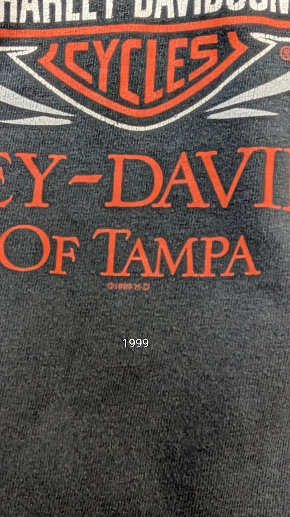 Close-up of a dark gray or black t-shirt fabric featuring orange and white Harley-Davidson branding and the text 'CY-DAVIDSON OF TAMPA' with '1999' printed below.