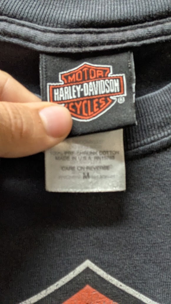 Close-up of a Harley-Davidson garment neck label being held by a finger, showing the brand patch and inner care tag on dark gray fabric.