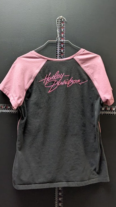 A Harley-Davidson raglan t-shirt hanging on a metal hanger against a dark gray wall. The shirt features a dark gray body and light pink sleeves, with the brand name 'Harley-Davidson' written in pink script across the upper back.