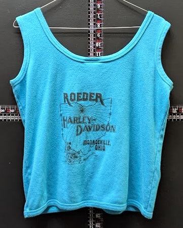 A bright turquoise blue cropped tank top with a Harley-Davidson logo graphic on the front, hanging on a wire hanger against a dark gray wall with a measuring tape visible behind it.