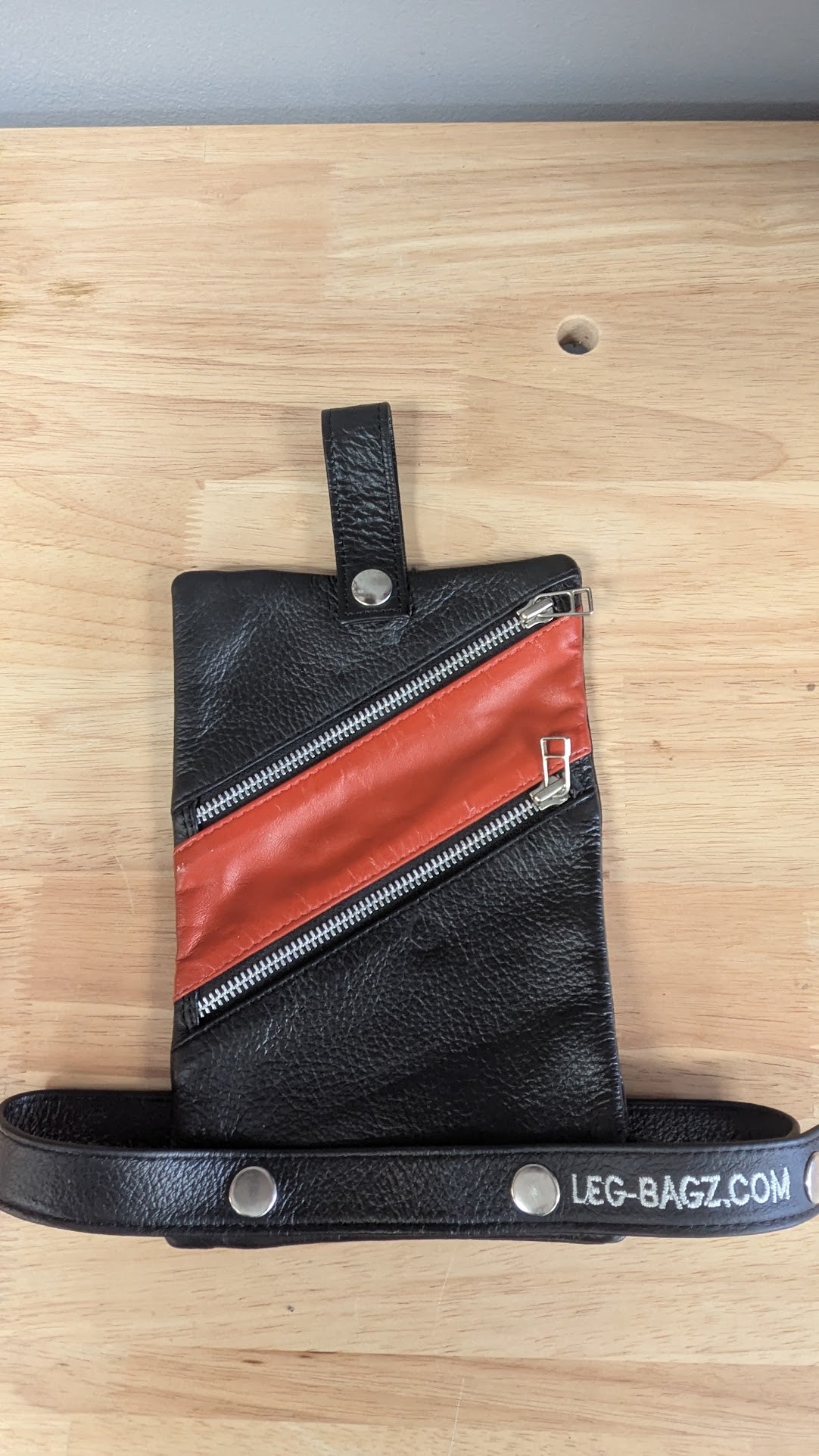 Black leather leg bag with diagonal orange-red accent panel and silver zipper, shown on a light wood surface with a black strap printed with LEG-BAGZ.COM.