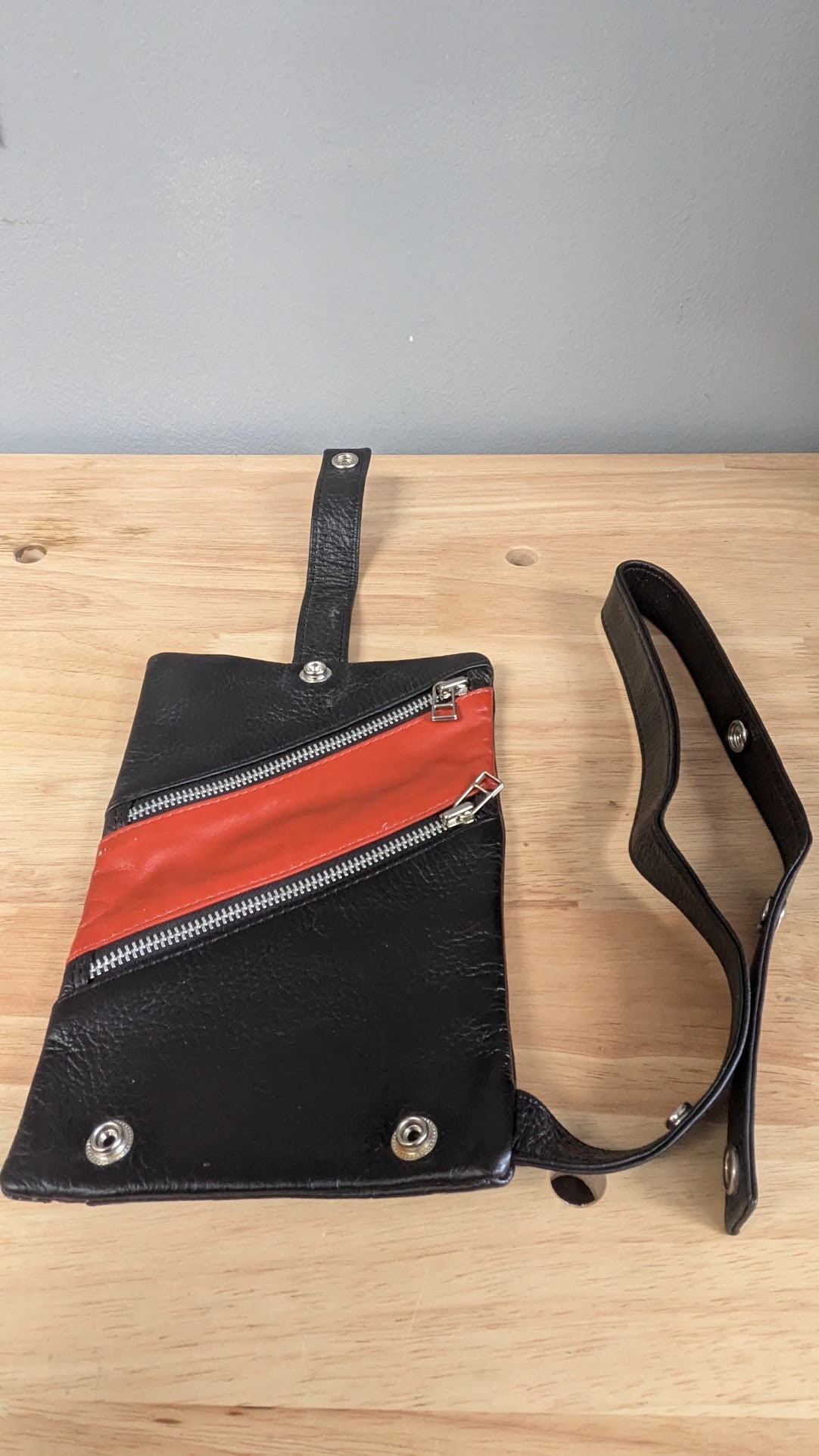 Black leather shoulder bag with a diagonal red leather stripe and silver zippers, displayed on a light wood surface against a gray wall.