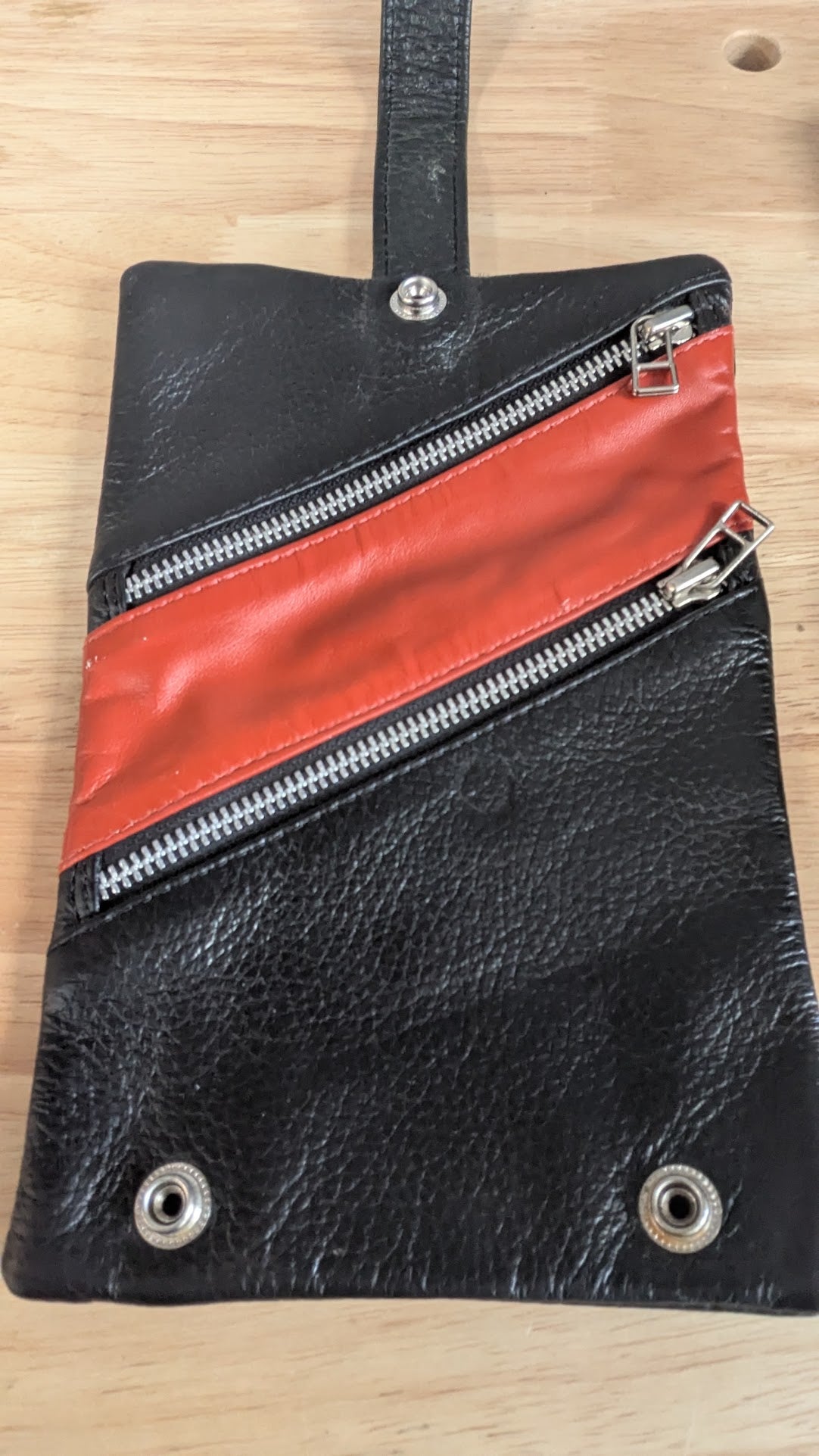 Black textured leather pouch with a diagonal red leather stripe and silver zippers, shown on a light wood background.
