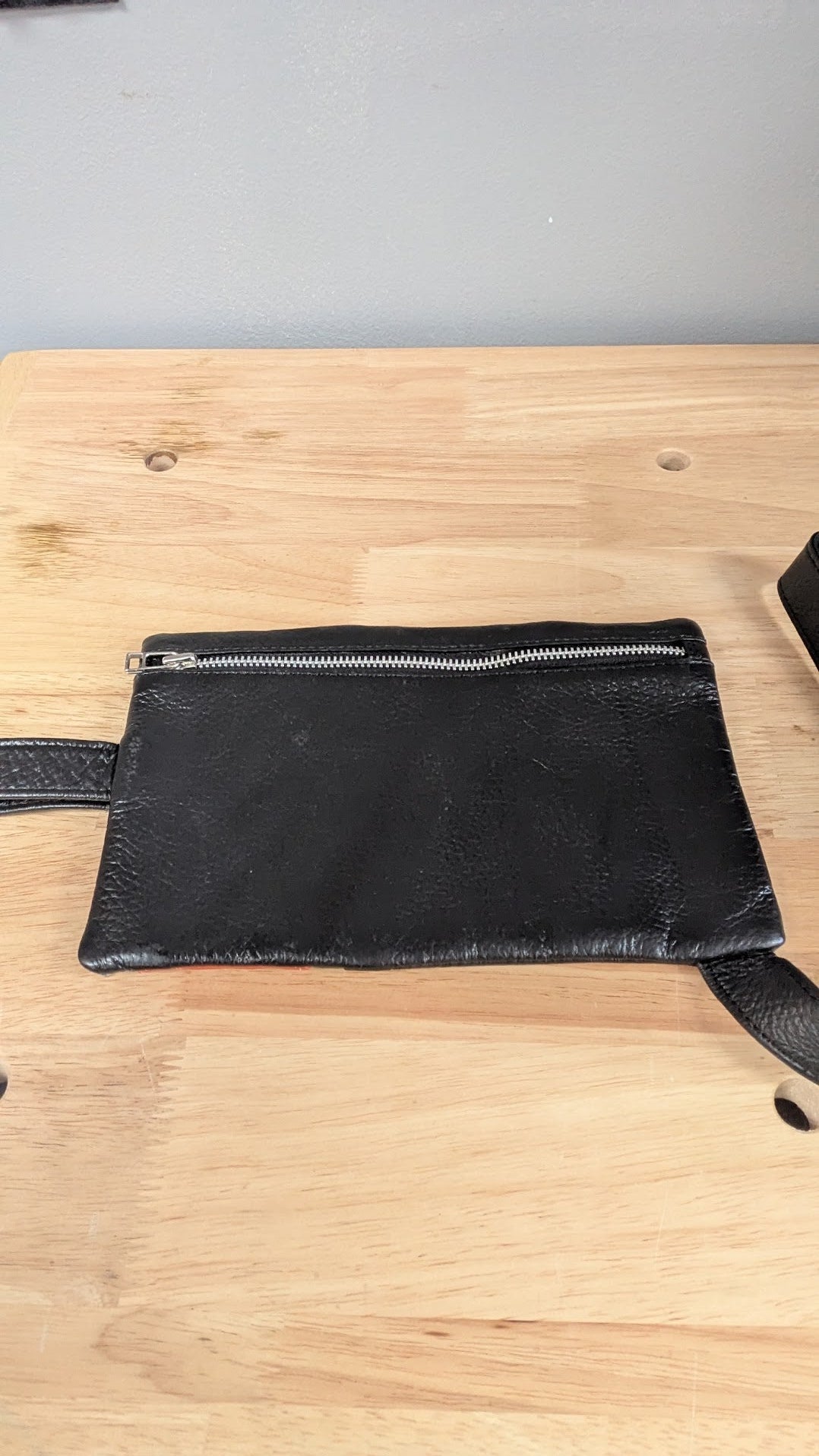 A black textured leather pouch with a silver zipper and strap, laid flat on a light wood tabletop.