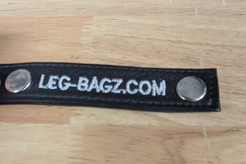 A close-up of a black leather strap with two silver magnetic snaps and white text reading 'LEG-BAGS.COM', held by a hand over a light wood grain surface.