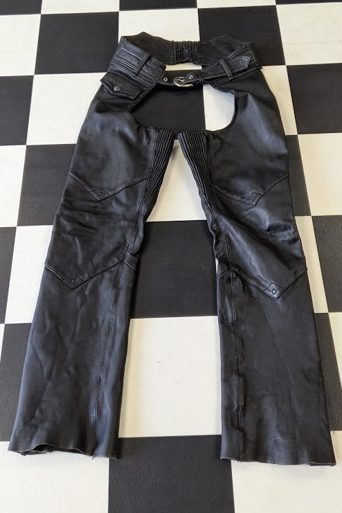 Black leather chaps pants with a belt loop waist and front zipper closure, laid flat on a black and white checkered floor.