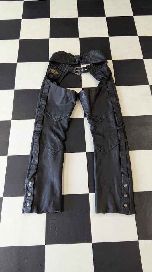 Black leather chaps with snap-button cuffs and an adjustable waist strap, laid flat on a black and white checkered floor.