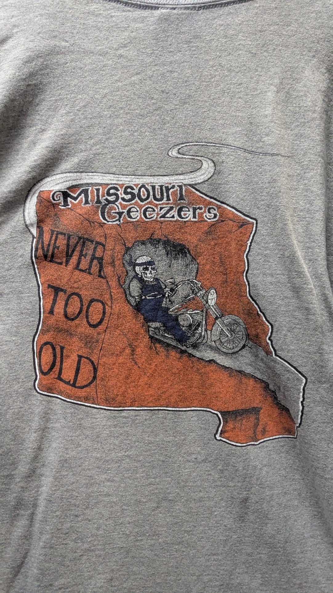 Close-up of a heather gray t-shirt featuring a distressed orange graphic of the state of Missouri with the text 'Missouri Geezers' and 'NEVER TOO OLD' alongside a cartoon skeleton riding a motorcycle.