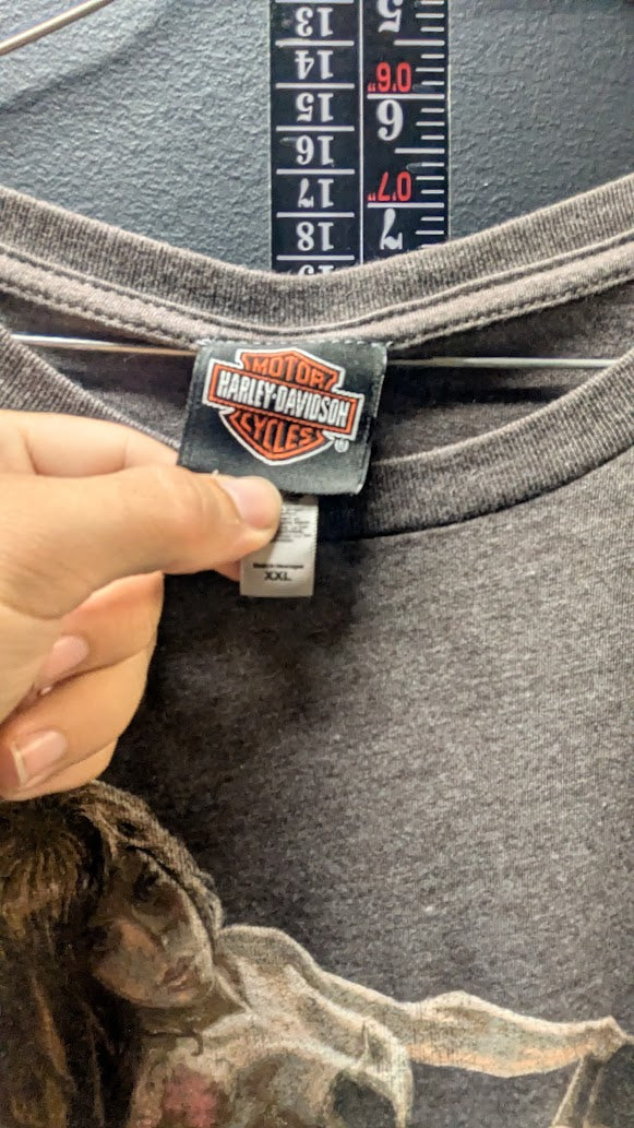 Close-up of a dark gray Harley-Davidson t-shirt with a hand holding the brand label, with a size measurement ruler visible behind the collar.