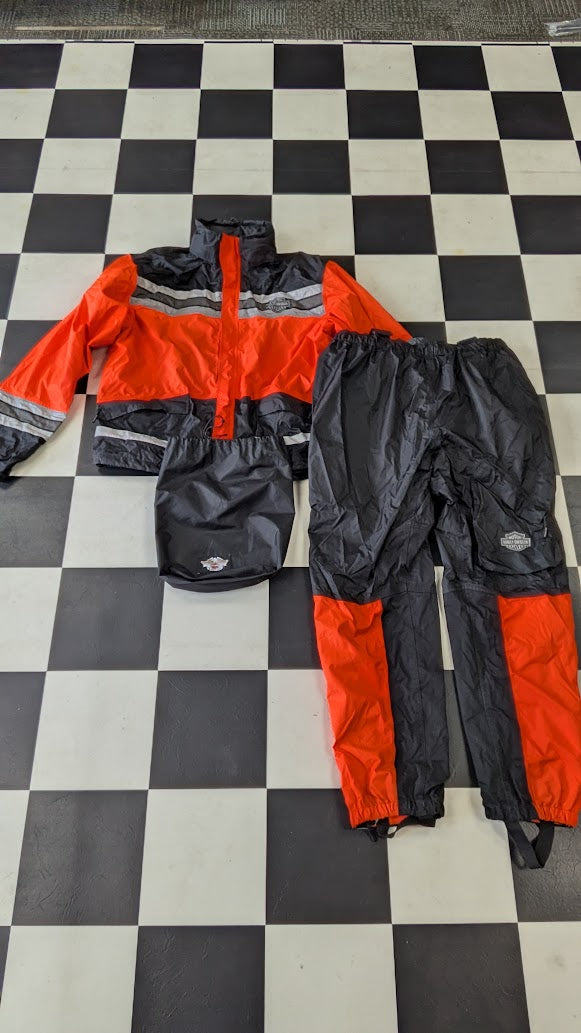 A three-piece rain suit laid flat on a black and white checkered floor, consisting of a hooded jacket, matching pants, and a small rain bag. The jacket is bright orange with dark gray/black sections and reflective silver stripes, while the pants are dark gray/black with orange sections on the lower legs.