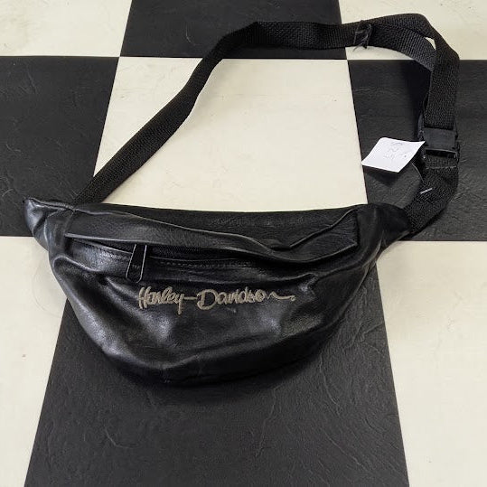 A black faux-leather fanny pack with a white script Harley-Davidson logo, resting on a black and white checkered floor.