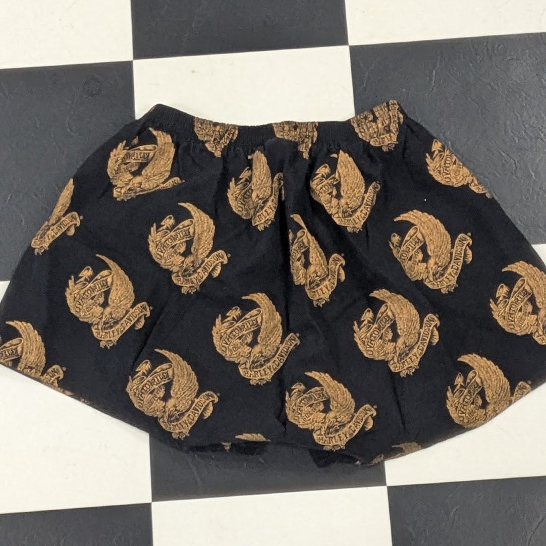 Black shorts with a repeating gold cartoon character print, laid flat on a black and white checkered floor.