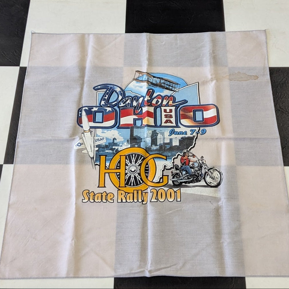 A square white fabric bandana or napkin featuring a colorful '2001 King State Rally' graphic, laid flat on a black and white checkered floor.