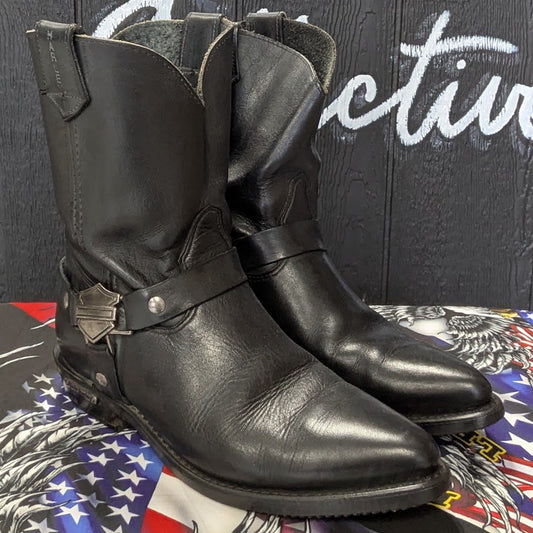 A pair of black leather biker boots with western-style stitching and a small metal accent, displayed on a graphic-patterned surface in front of a dark wood sign that reads 'Biker Collective'.