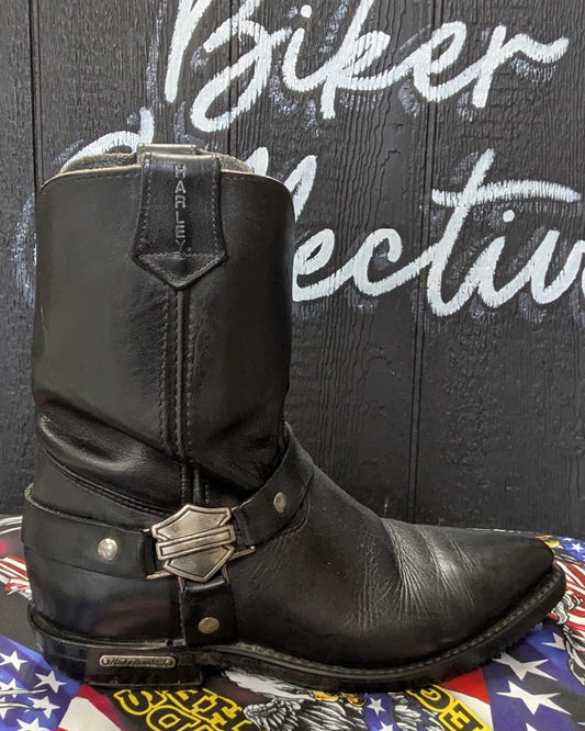 A single black leather motorcycle boot with silver hardware, including a Harley-Davidson bar-and-slot buckle, displayed on a graphic mat against a dark wood wall with white painted text.